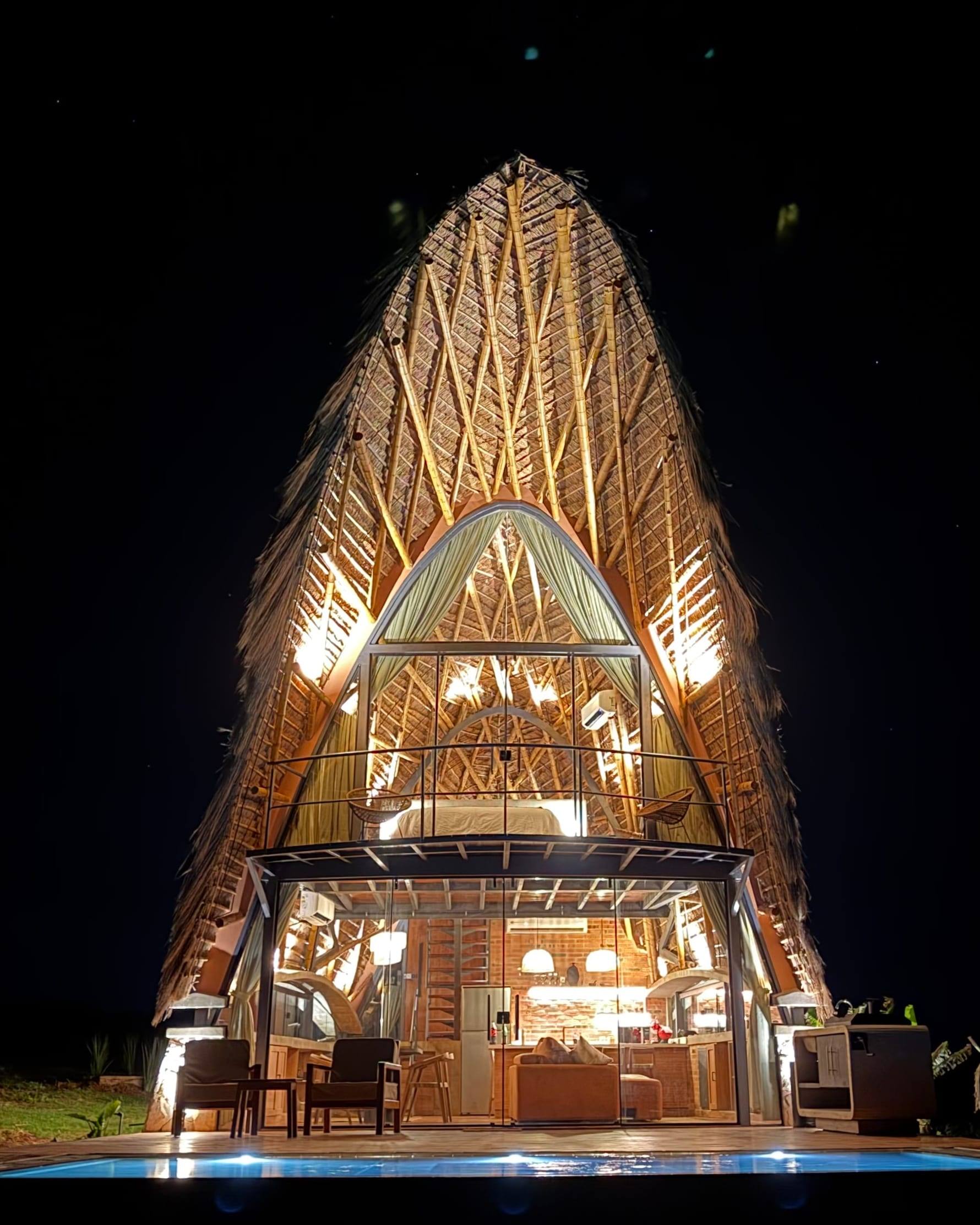 El bungalow de noche, toda la estructura de tacuara iluminada desde adentro y la piscina encendida al pie.